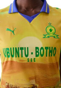 Maglia da calcio gialla con colletto blu, caratterizzata dalla scritta "UBUNTU-BOTHO" in verde, una grafica del sole e un logo Puma in ricamo verde.