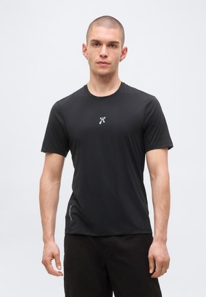 Mladić s kratkom kosom nosi jednobojnu crnu majicu kratkih rukava i crne hlače, stoji na jednobojnoj svijetloj pozadini.