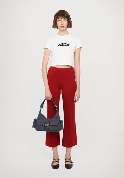 Νεαρή γυναίκα με λευκό κοντό μπλουζάκι που έχει την ένδειξη "Icon", κόκκινες κοντές παντελόνες, κρατώντας μια γκρίζα τσάντα και φορώντας μαύρα παπούτσια με λουράκια.