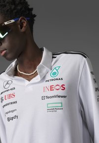 White polo shirt with black stripes on shoulders, featuring multiple logos in red and black. Accessories include a layered pearl necklace and sunglasses.