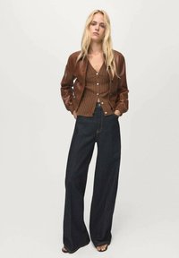 Giacca bomber in pelle marrone sovrapposta a un cardigan marrone a coste, abbinata a jeans scuri a gamba larga. Design semplice con tonalità neutre.