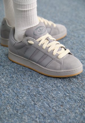 Szare trampki Adidas Campus z kremowymi sznurowadłami, noszone z białymi skarpetkami na niebieskim wykładanym dywanie.