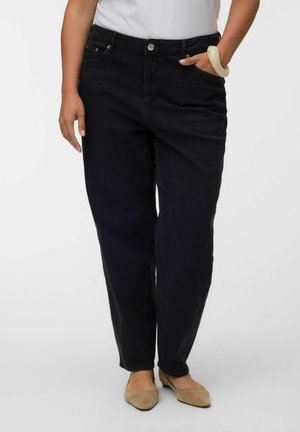 Femme portant un jean noir taille haute à jambes droites, un haut blanc, des ballerines beiges et un grand bracelet blanc à son poignet droit.