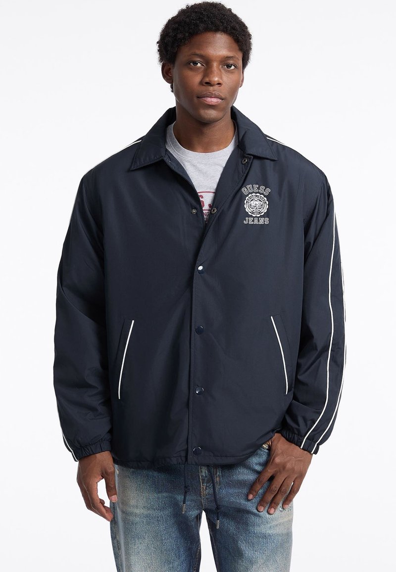 Marineblauer Windbreaker mit weißen Streifen, Druckknopfverschluss und gesticktem Logo. Verfügt über elastische Bündchen und seitliche Taschen.