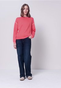 Femme debout devant un fond pâle, portant un pull corail à manches longues, un jean large bleu foncé et des chaussures de couleur claire, une main dans la poche.