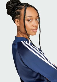 Navy blue athletic jacket featuring white stripes on sleeves, made of smooth fabric with a stand-up collar and decorative braided hair.