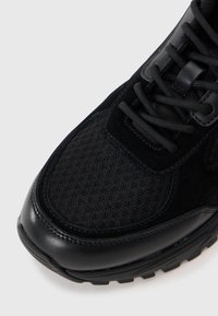 Black sneaker featuring a mix of textured suede and mesh materials, with a smooth leather toe cap and black laces. Rubber outsole.