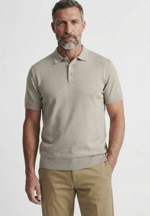 Homme d'âge moyen avec une barbe grise portant un polo beige à manches courtes et un pantalon kaki, debout devant un fond clair uni.