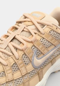 Baskets en mesh beige avec des superpositions de tissu croisées, des accents texturés, un swoosh gris clair et des lacets plats pour un ajustement sécurisé.