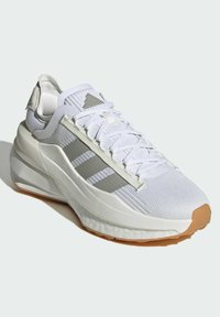 Zapatilla atlética blanca con parte superior de malla, detalles grises, suela texturizada y diseño de tres franjas. Presenta un talón acolchado para mayor soporte.