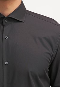 Zwart overhemd met knopen, voorzien van een moderne kraag, een gladde textuur en drie zichtbare knopen. De stof lijkt lichtgewicht en kreukbestendig.