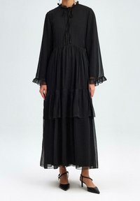 Robe noire à volants avec des manches longues, des accents froncés aux poignets et à l'ourlet, et un col noué, fabriquée en tissu léger pour une apparence fluide.