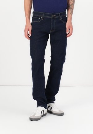 Homme portant un jean droit bleu foncé et des baskets blanches avec des bandes noires, debout devant un fond uni.