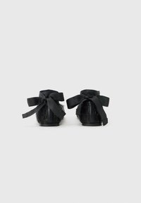 Chaussures noires avec de grands nœuds en tissu à l'arrière, matériau texturé avec des embellissements, et un design à semelle plate.