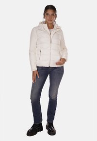 Giacca imbottita bianca con cappuccio, dotata di una zip frontale e tasche laterali, abbinata a jeans blu scuro e stivali neri stringati.