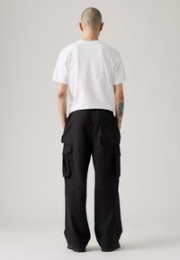 Mand med glemt hoved iført hvid t-shirt og løse sorte cargo bukser, stående med ryggen mod en lys ensfarvet baggrund.