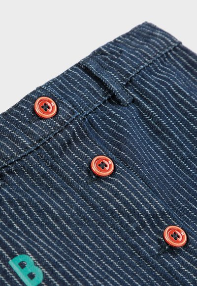 Prugasta plava denimska tkanina sa dva crvena dugmeta s crnim cvjetnim uzorcima. Prednji panel uključuje slovo "B" u tirkiznoj boji.