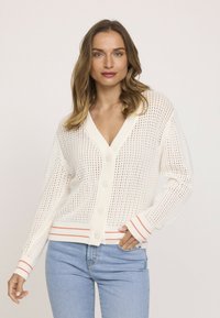 Cardigan en maille blanche avec un design ajouré, col en V, fermeture à boutons sur le devant, et accents rayés rouges sur les poignets et l'ourlet. Associé à un jean bleu.