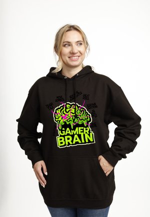 Femme souriante portant un sweat à capuche noir avec un graphique de cerveau vert néon étiqueté "GAMER BRAIN" et des termes de gaming comme "Combos de mouvements", "Codes de triche" et "Lieux de réapparition".