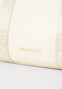 Kremfarget lærhåndveske med en teksturert overflate, innpreget merkevare "VALENTINO," og en bred logo-trykt stoffstropp.