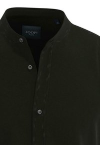 Ciemnozielona koszula z stojącym kołnierzykiem, z patką zapinaną na guziki i fakturowaną tkaniną. Etykieta marki widoczna wewnątrz kołnierza.