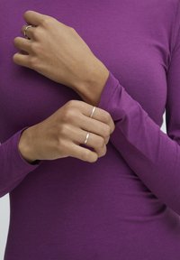 Långärmad, åtsittande lila topp med jämn textur; två silvriga ringar på fingrarna. Närbild av händer som korsar varandra vid handleden.