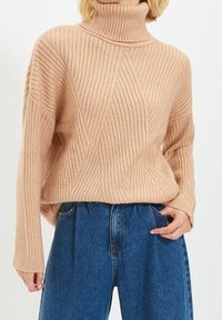 Jasno beżowy sweter z wysokim kołnierzem, o swobodnym kroju i pionowym wzorze. W komplecie z niebieskimi, wysokogatunkowymi dżinsami.
