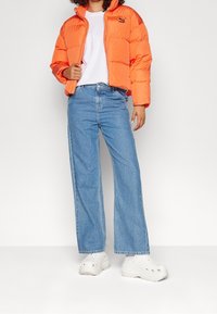 Giacca arancione imbottita con zip, abbinata a jeans a gamba larga blu chiaro e scarpe bianche senza lacci. La giacca presenta un logo sul petto.