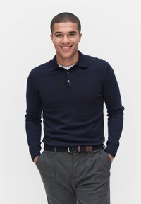 Tummansininen pitkähihainen polo pusero, jossa on perinteinen kaulus ja kolme nappia. Yhdistetty harmaisiin räätälöityihin housuihin ja teksturoituun vyöhön.