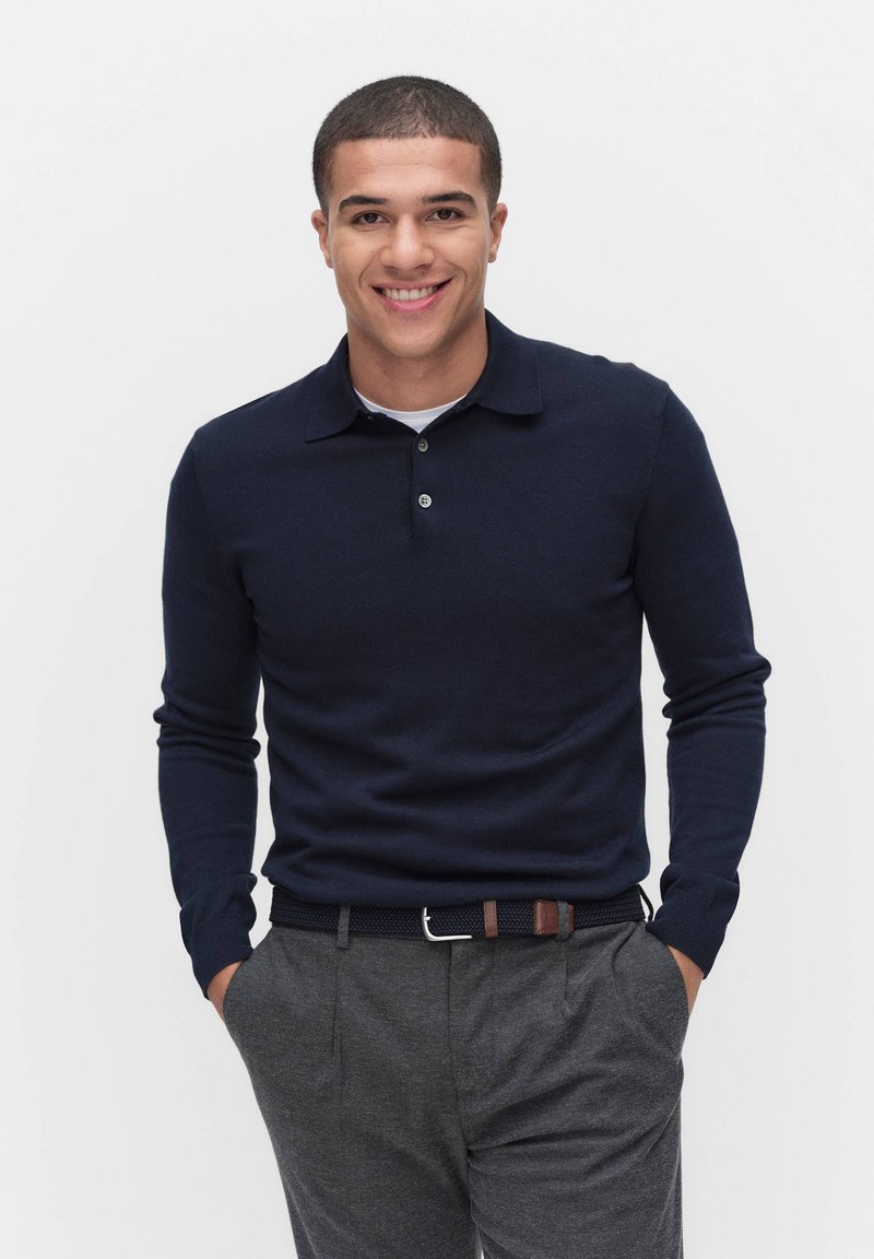 Tummansininen pitkähihainen polo pusero, jossa on perinteinen kaulus ja kolme nappia. Yhdistetty harmaisiin räätälöityihin housuihin ja teksturoituun vyöhön.