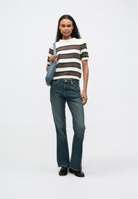 Gestreifter Strickpullover in Creme und Braun, blaue Schultertasche und weit geschnittene blaue Jeans. Das Modell steht vor einem schlichten Hintergrund.