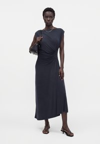 Femme portant une robe midi noire sans manches froncée, tenant un sac à main noir sur une épaule, portant des sandales noires à petits talons.
