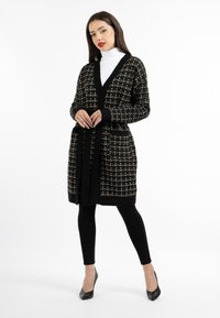 Cardigan testurizzato nero e oro con scollatura a V, maniche lunghe e cintura in vita, abbinato a leggings neri e tacchi alti.
