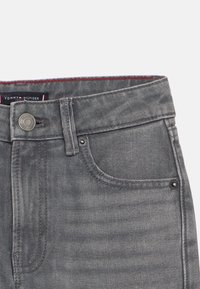 Jeans in denim grigio con chiusura a bottone, diverse tasche e un accento rosso all'interno della cintura. Etichetta del marchio visibile.