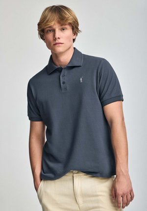 Polo de color gris oscuro con cuello, tapeta de tres botones y logo bordado en el pecho; combinado con pantalones de color claro.