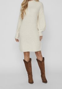 Robe-pull crème en maille avec manches longues ballon et col montant. Portée avec des cuissardes en daim marron au sommet pointu.