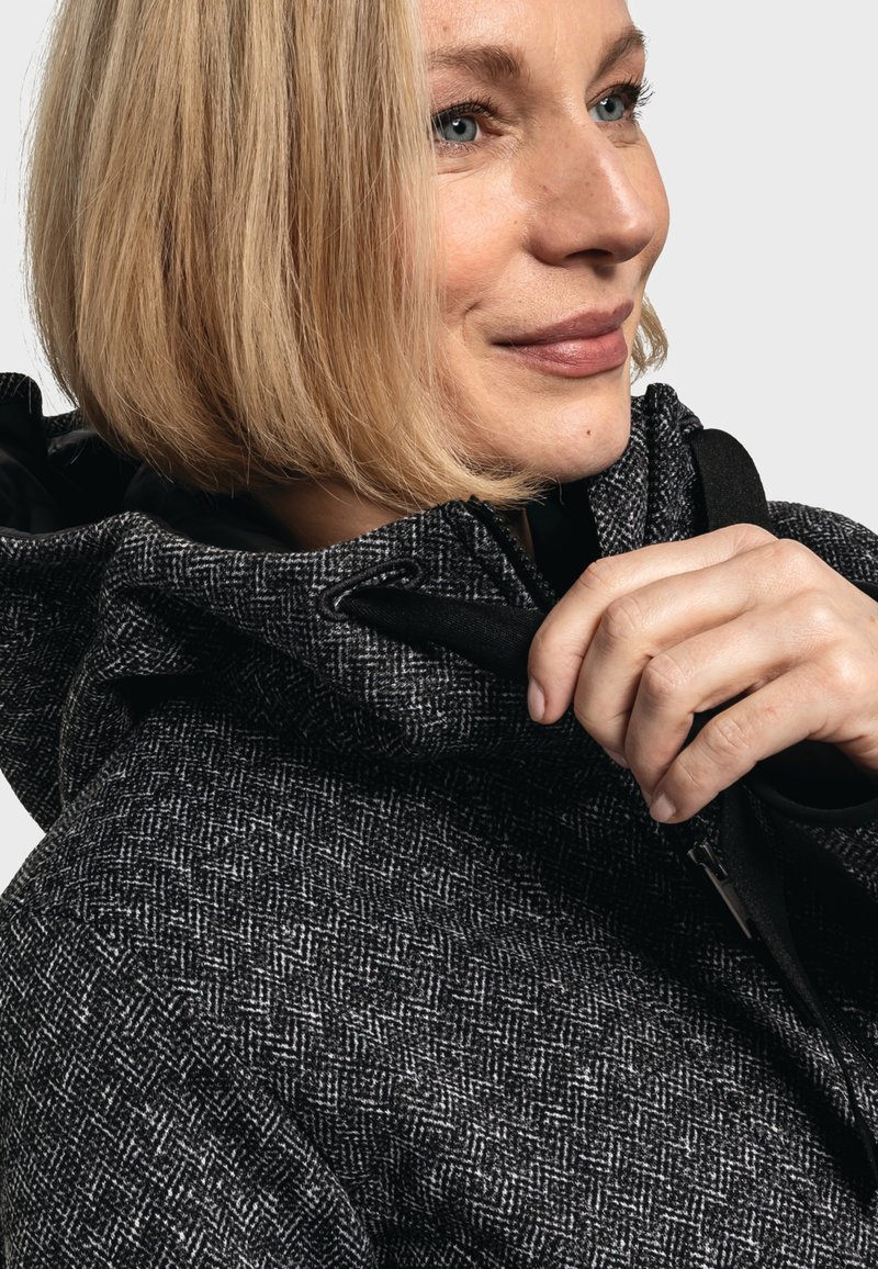 Giacca a motivi a spina di pesce bianco e nero con colletto alto, realizzata in un tessuto texture e dotata di cappuccio regolabile, con zip e dettagli neri.