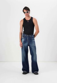 Ung man som bär vita smala solglasögon, svart ärmlös topp, vida blå jeans och svarta skor, stående mot vit bakgrund.