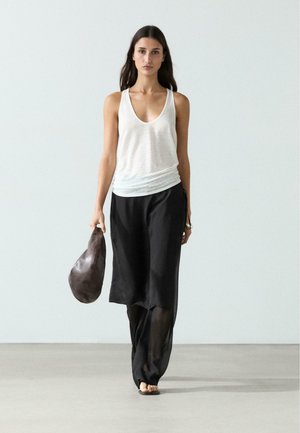 Kvinde går i sorte bukser med vide ben, hvid ærmeløs top, sorte sandaler og bærer en mørkebrun lædertaske i et minimalistisk miljø.