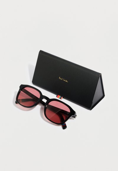 Černé obdélníkové sluneční brýle s červenými čočkami, robustním rámem a stříbrnými detaily. Dodávají se s černým trojúhelníkovým pouzdrem označeným "Paul Smith."