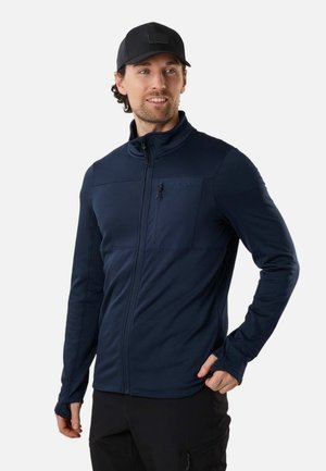 Mężczyzna w granatowej kurtce zapinanej na zamek z otworami na kciuki i czarnej czapce, stojący z jedną ręką w kieszeni, patrzący w bok.