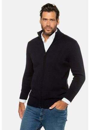 Maglione zip con cerniera di colore navy realizzato in tessuto a maglia, con colletto e polsini a costine, che presenta una zip frontale e un design aderente. Indossato sopra una camicia bianca.