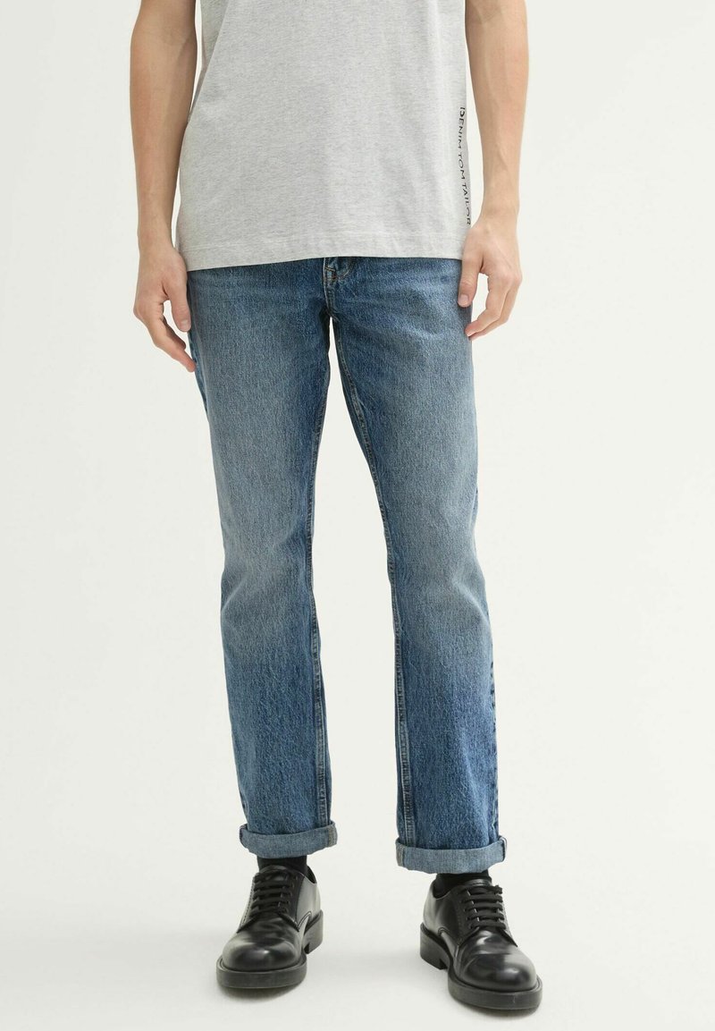 Jeans de mezclilla azul con un corte recto, lavado desgastado y puños enrollados; combinados con zapatos de cuero negros y una camiseta gris claro.