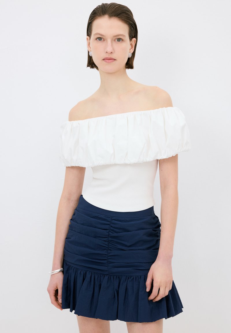Top bianco off-the-shoulder con scollo a volant, abbinato a una mini gonna arricciata blu scuro, con fondo svasato e tessuto texturizzato.