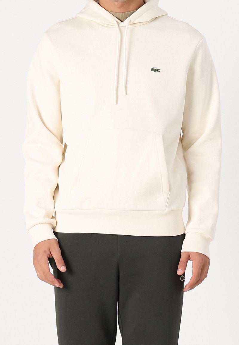 Homme portant un sweat à capuche crème avec poche avant et cordons, associé à un pantalon foncé montrant un petit logo sur la cuisse gauche.