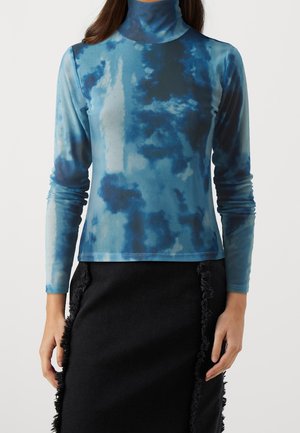 Kobieta w niebieskim golfie typu tie-dye z długim rękawem i czarnej spódnicy z frędzlami na bocznych szwach.
