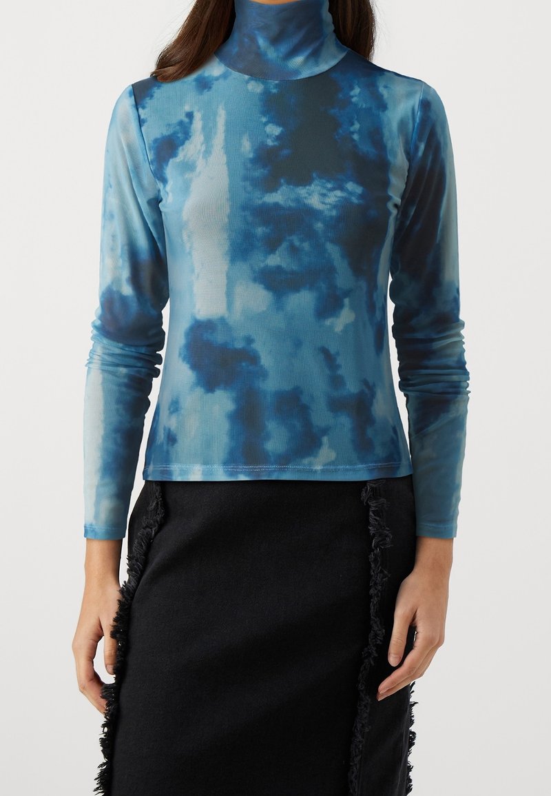 Femme portant un col roulé à manches longues tie-dye bleu et une jupe noire avec des coutures latérales à franges.