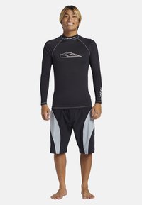 Černá surfová košile s dlouhým rukávem a bílým logem, kombinovaná s černými a šedými plážovými kraťasy s zakřivenými bočními pruhy. Naboso.