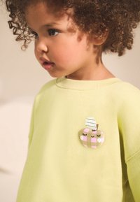 Light yellow sweatshirt featuring a heart-shaped patch with a plaid apple design, adorned with a striped leaf and embroidered facial details.