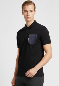 Černé polo tričko s límečkem, s tmavým denimovým kapsičkou na levé straně hrudi. Krátké rukávy a hladká texturovaná látka.